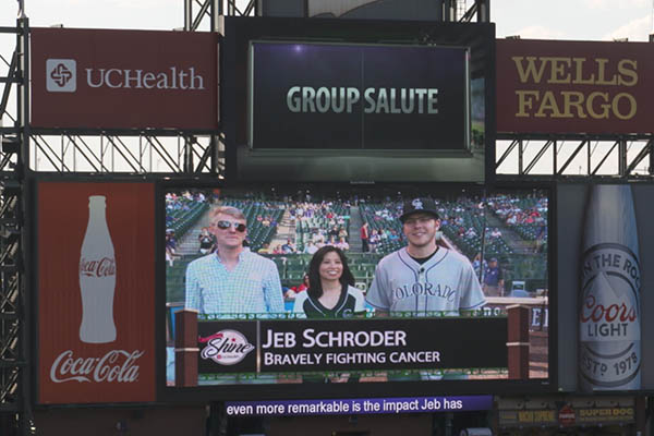 A sign at Coors Field shows Jeb Schroder with his doctors during a Rockies game. The scoreboard says, "Jeb Schroder: Bravely Fighting Cancer."