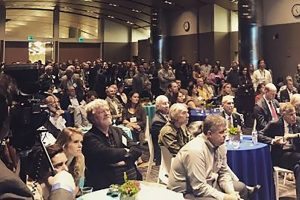 A large group of people sit in a conference room listening to a speaker.