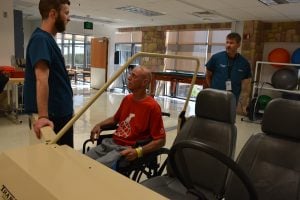Patrick Buxton, a patient in the Rehabilitation Patient Care Unit at Memorial Hospital Central, works with Andrew Bettlach, an occupational therapist, to practice transferring from a wheelchair into a simulated car. Gregory Braeger, therapy manager, looks on.