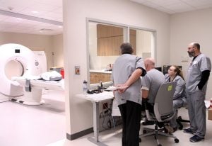 A team of imaging experts at UCHealth Longs Peak Hospital practice using a new CT machine during a training. You see the workers in one room and the large white CT machine in an adjacent room.