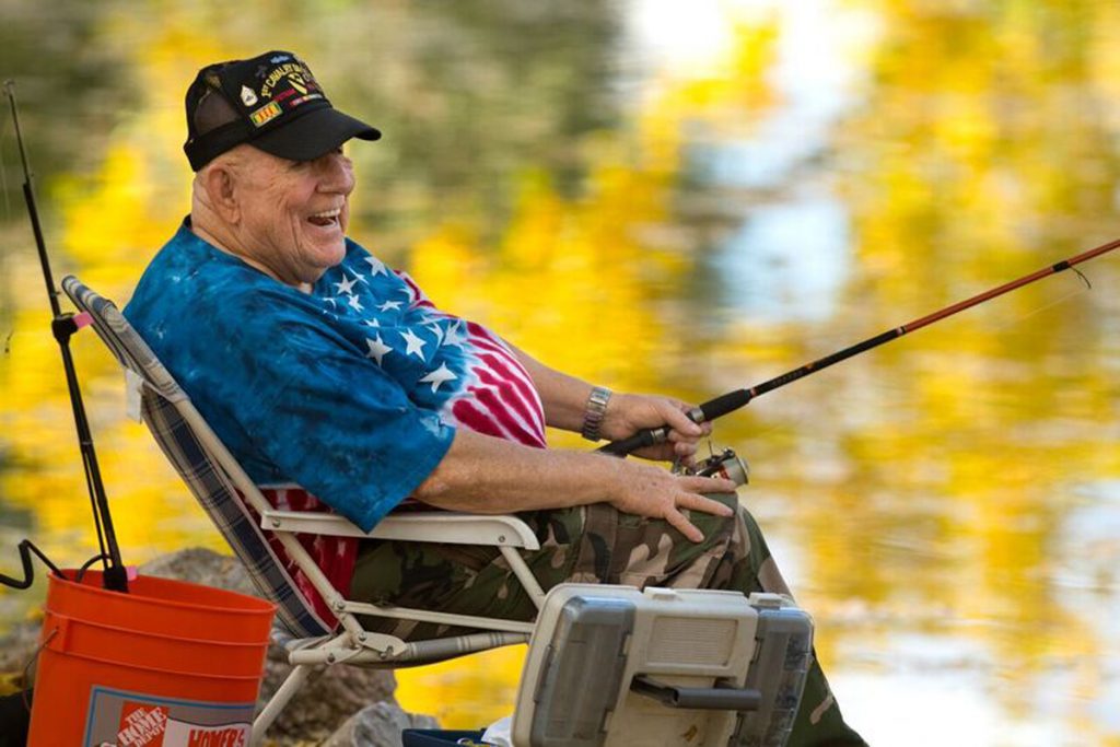 J.D. Hill sitting in a chair fishing by a lake with a tie-dye american flag shirt