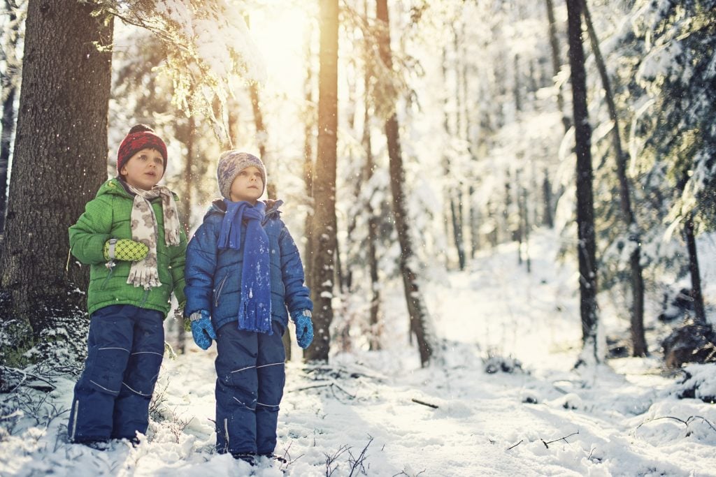 Two children wear winter parkas and hats as they stand in a snowy forest.