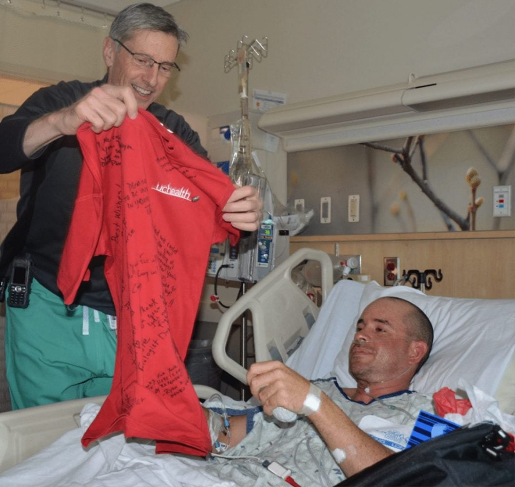 Dr. Daniel Huddle stands next to a patient's bedside and gives him a red T-shirt.