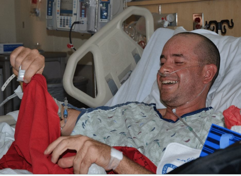 A patient in a hospital bed smiles after receiving a red T-shirt signed by his caregivers.