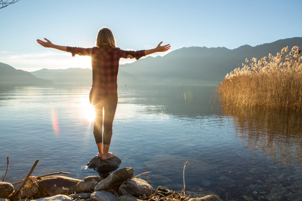 A woman balances on a rock while overlooking a lake.
