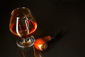 Whiskey glass and smoking pipe on black background. Cognac glass. Brandy glassful. Cognac france. Scotch drink. Smoking pipe