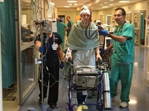 A photo of a man walking in the hospital with caregivers and life-saving medical devices nearby.