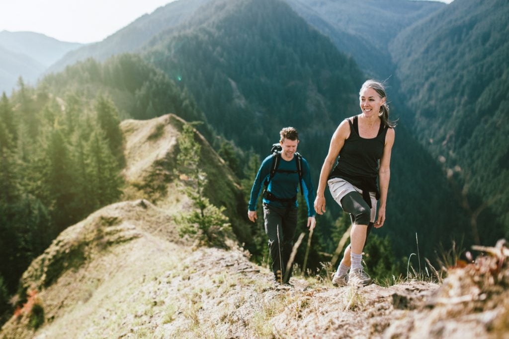 A fit man and woman in their 50's hiking the trail on a mountain ridge, a beautiful river gorge spreads out behind them. They smile, enjoying the exercise, the beauty of nature, and staying strong.