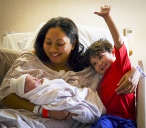 A mother, in the hospital bed, introduces her new-born son to her three-year-old son when the new-born son is only hours old. The three -year-old is ecstatic to have a new baby brother.