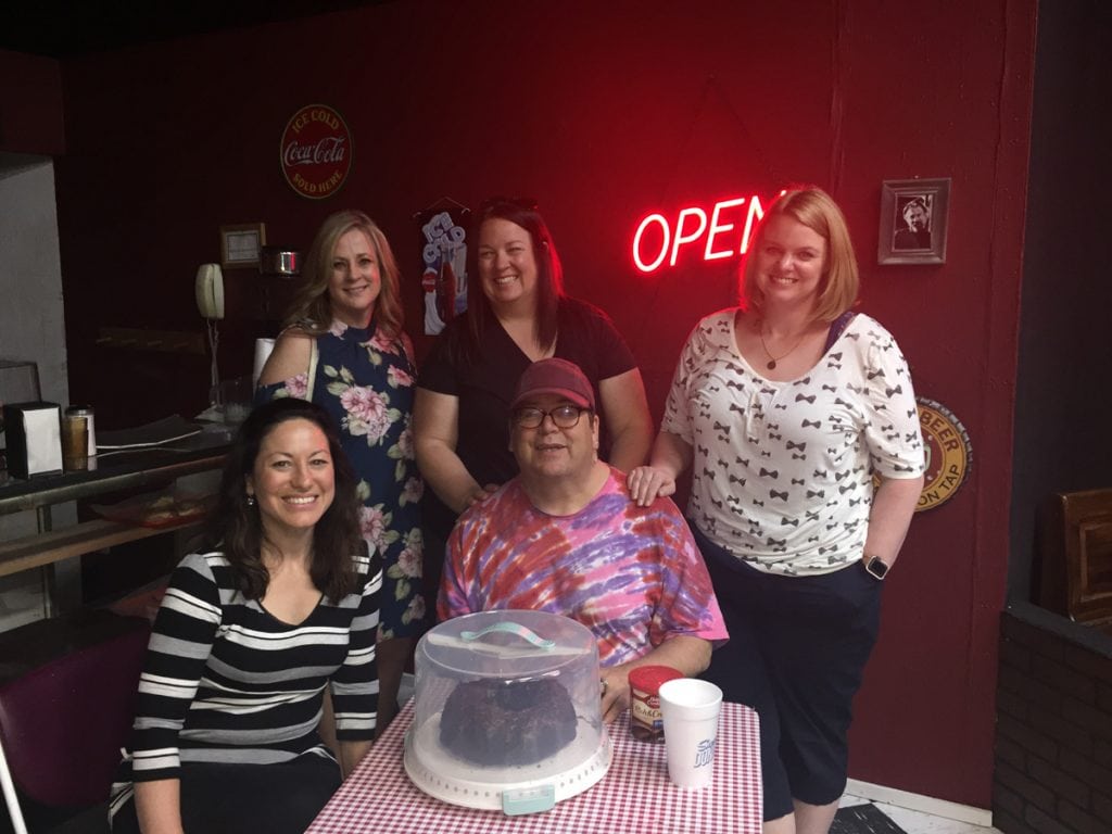Greg West and his health care team pose on the set for the play, Superior Donuts. West is thrilled to be back on stage afte rnearly dying from heart failure. They sit around a table with a sign that says "Open" behind them.