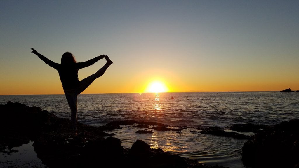 Louisa Drouet holds her leg out to her left as she does a yoga pose on a California beach at sunset.