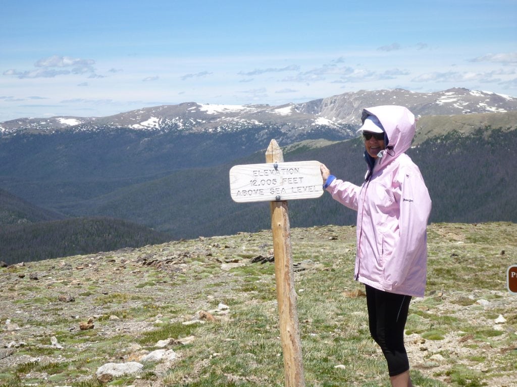 Trisha Poses in the summer on a high-altitude pass with a sign that says 12,000 feet above sea level.