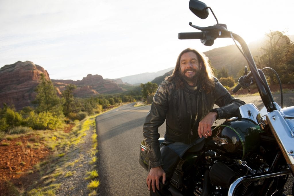 A young man with long hair poses on his Harley with the red rocks of Sedona in the background.