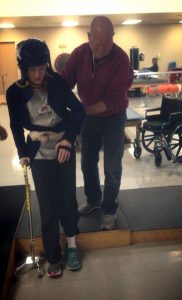 image of amy, which a helmet and a cane, being helped to walk by her father while in rehab.