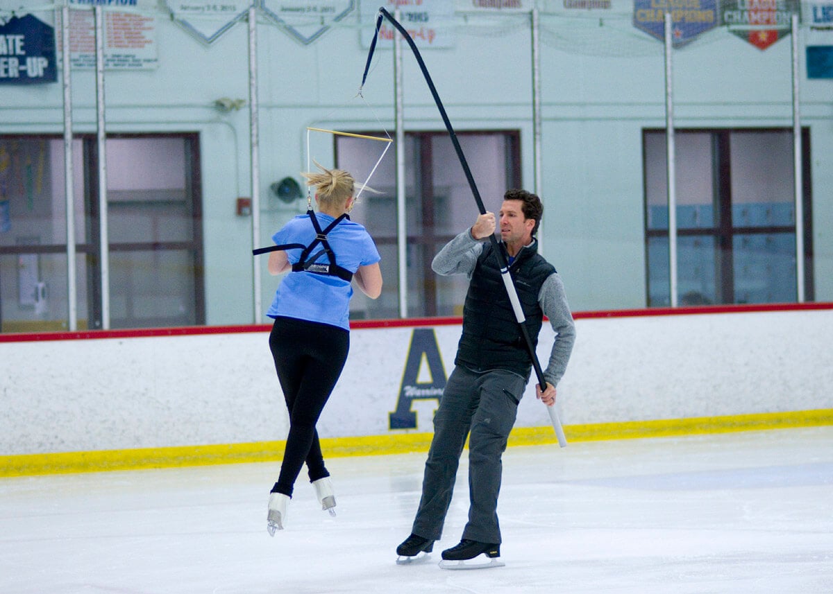skater turning in air while coach holds a harness to support her landing.