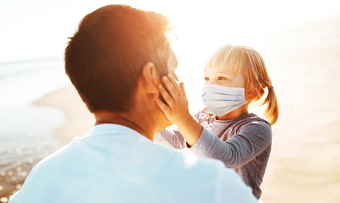 Little girl wears a mask and she cups her dad's face at the beach in this article about if it is safe to swim during covid..