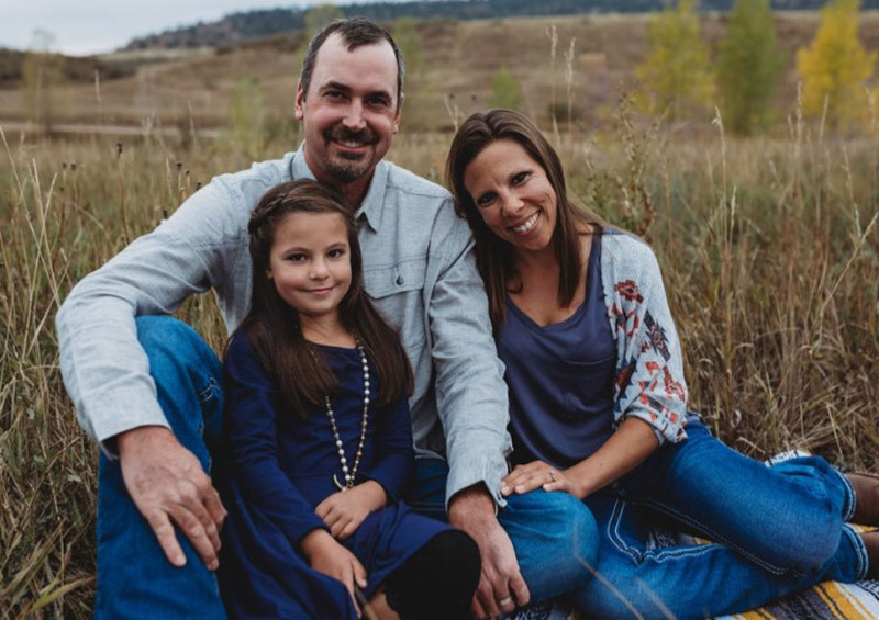 father, mom and daughter pose in a field for a family photo.