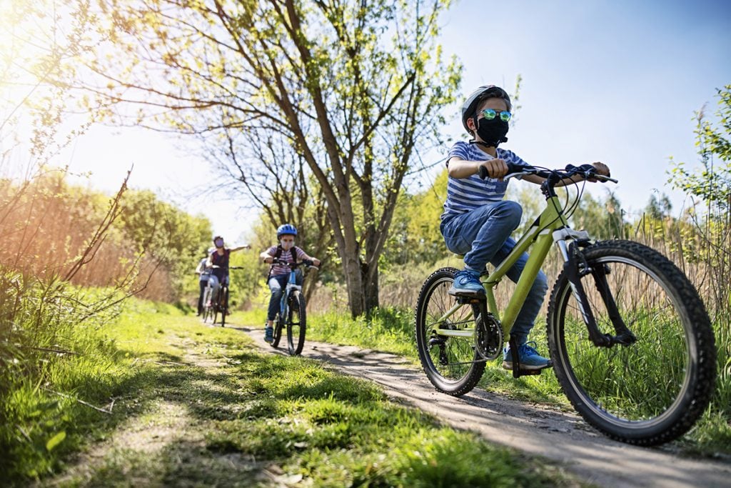 Kids ride bikes outdoors while wearing masks and practicing social distancing, demonstrating two ways to keep kids safe during this COVID-19 summer..