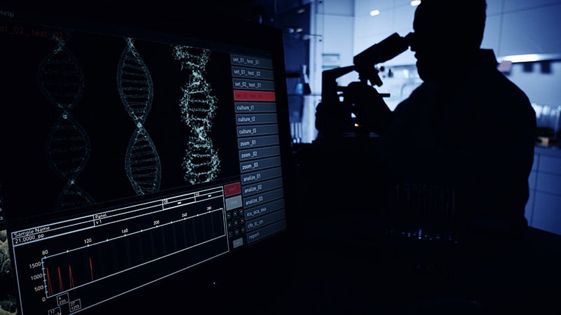 man looking into a microscope at coronavirus mutation, while DNA shows on the screen.