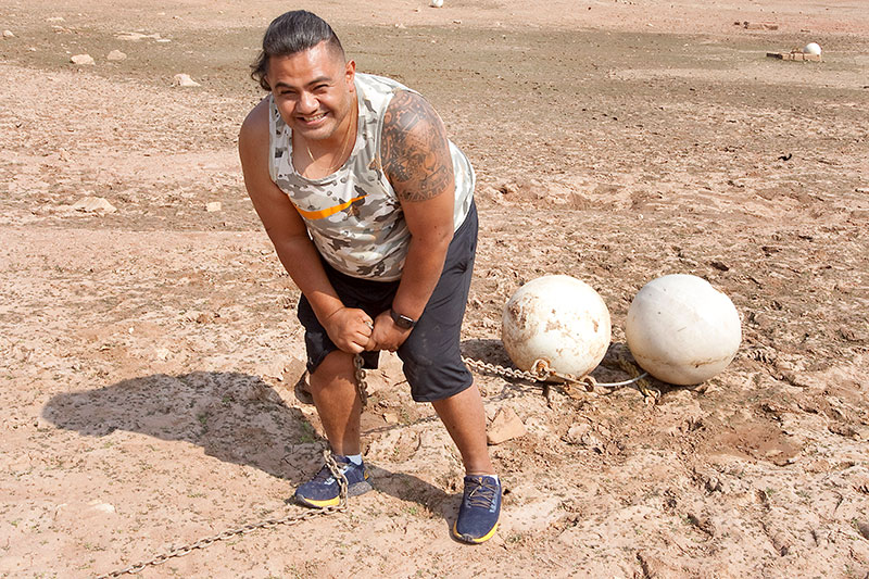 Edgar pulling two large balls on chains on a beach with a grin on his face.