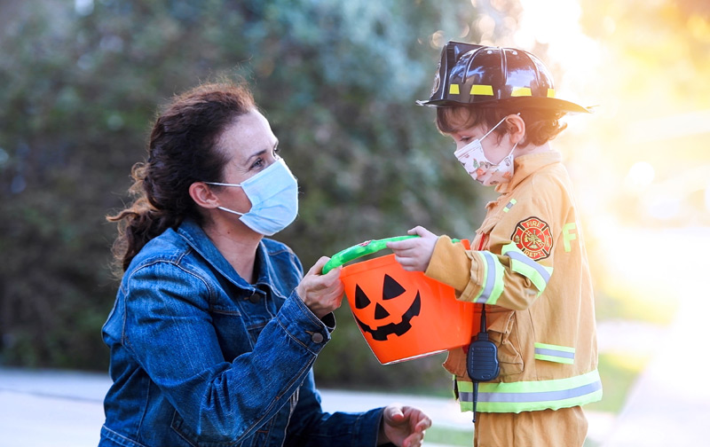 boy in a firefighter costume wearing a mask with his mother, also wearing a mask. Both demonstrating how to do halloween safely during COVID.