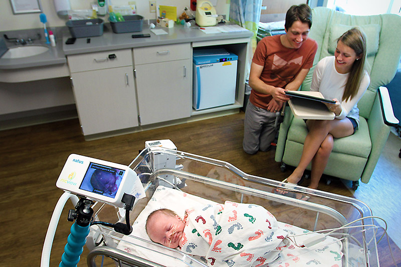 a NICU bedside camera points at baby Mason while his parents demonstrate how they can see him on their tablet.
